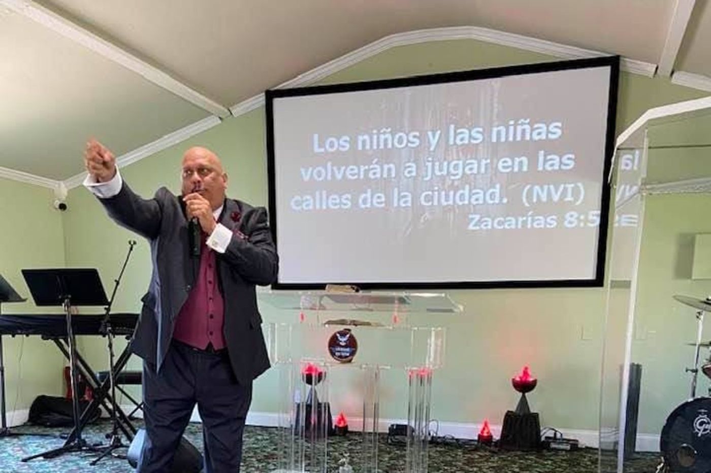 Man in dark suit speaking at podium with microphone beside projection screen displaying Spanish biblical text