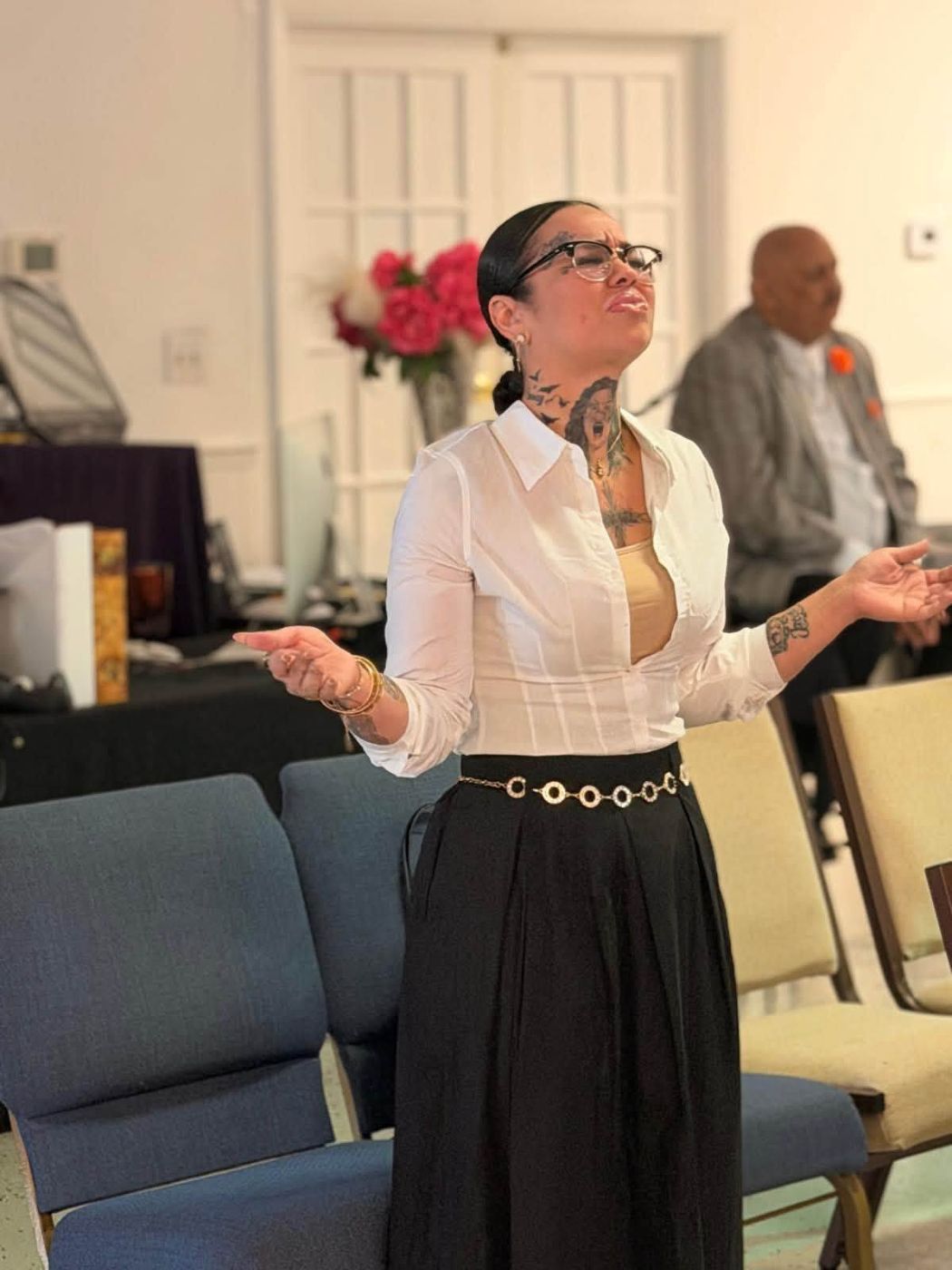 Woman with glasses and tattoos speaking at indoor event with flowers and attendees visible