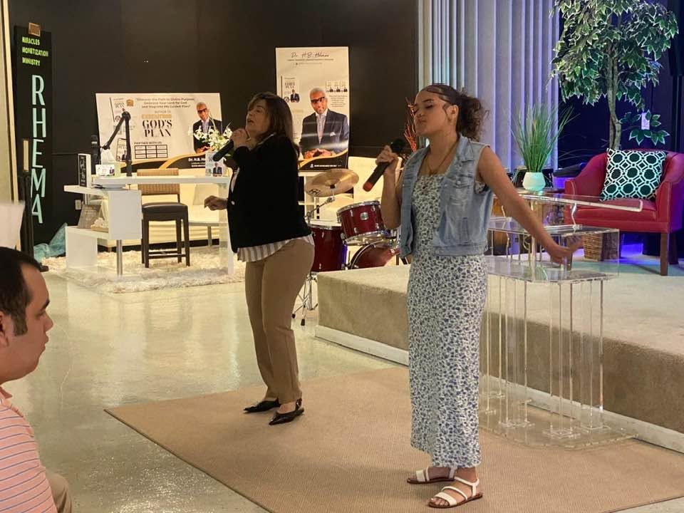 Two women singing with microphones at an indoor event venue with stage setup and audience seating