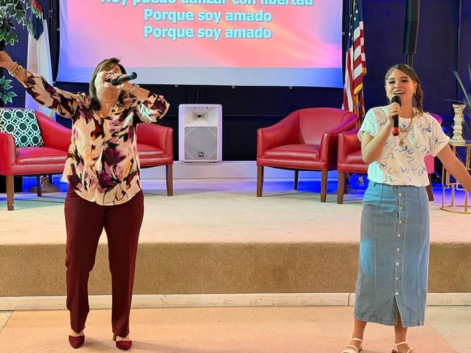 Two women singing on stage with microphones, red chairs, and Spanish text displayed on screen behind them