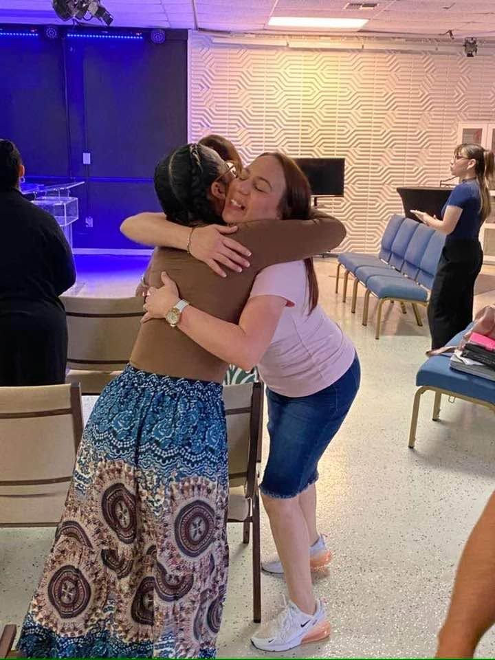 Two women embracing warmly in an indoor venue with blue and gold lighting and geometric wall panels
